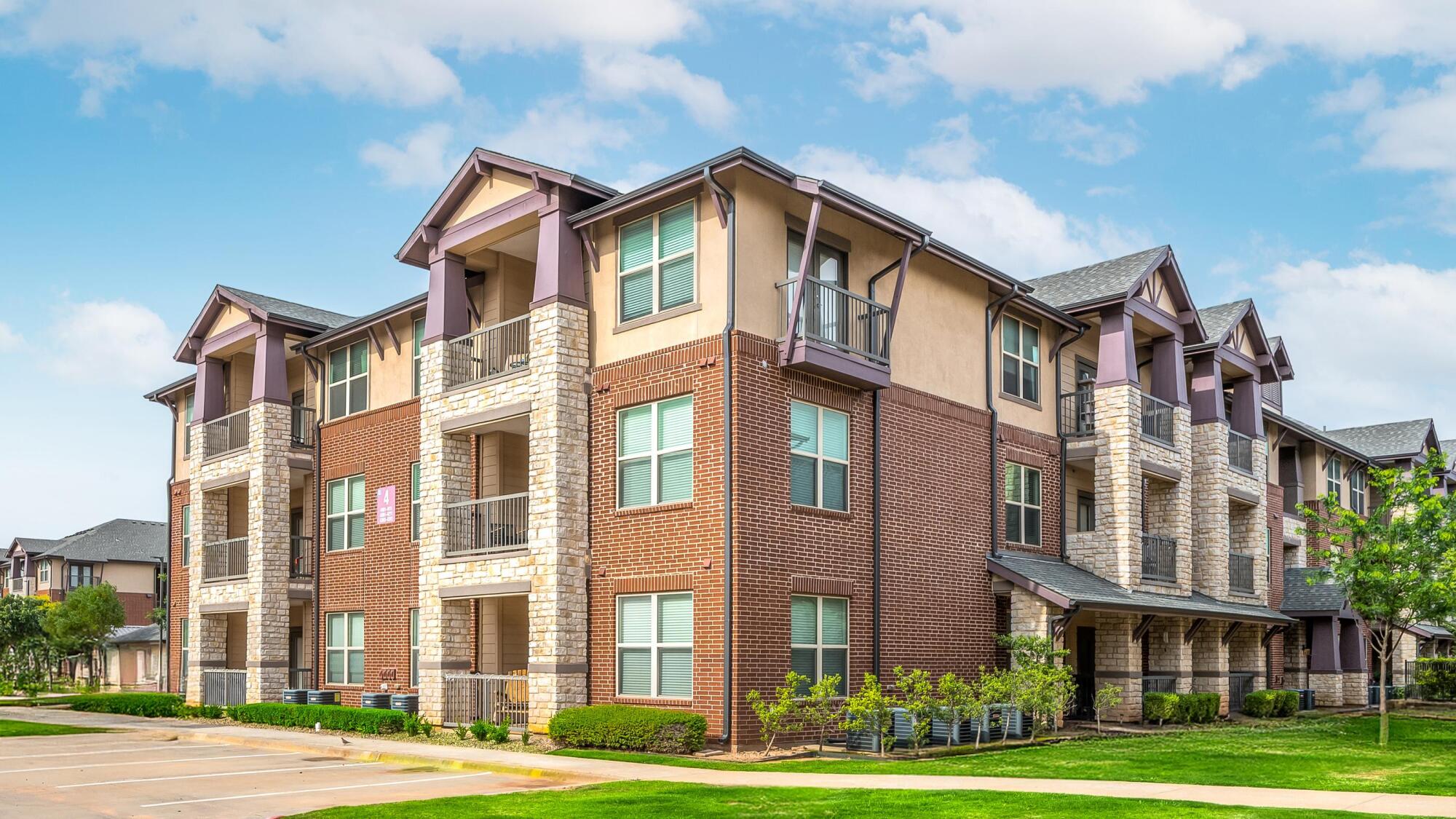 Modern three-story apartment building with balconies, brick and stone exterior, and green lawn under a blue sky.